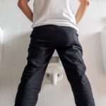 Homem sentado no vaso sanitário, com calça abaixada, representando dificuldade ou desconforto para urinar.