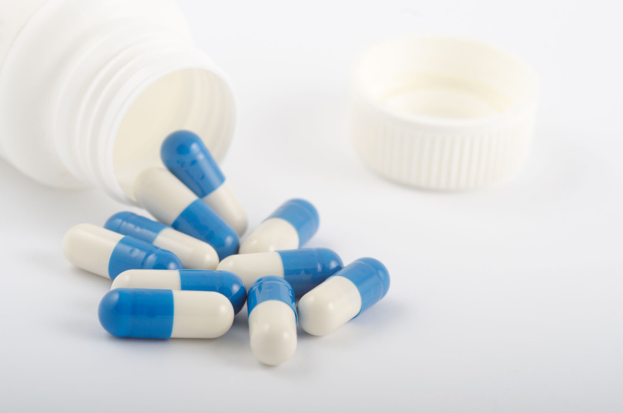 A imagem em close-up mostra várias cápsulas azuis e brancas espalhadas sobre uma superfície clara, tendo saído de um frasco branco tombado. As cápsulas estão relacionadas ao texto de blog “dutasterida para que serve”. A cena é uma representação limpa e clara de medicamentos e saúde. 