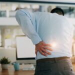 A imagem retrata um homem, presumivelmente um profissional de escritório, sofrendo de dor nas costas, o que pode estar relacionado ao tema próstata aumentada causa dor nas costas.